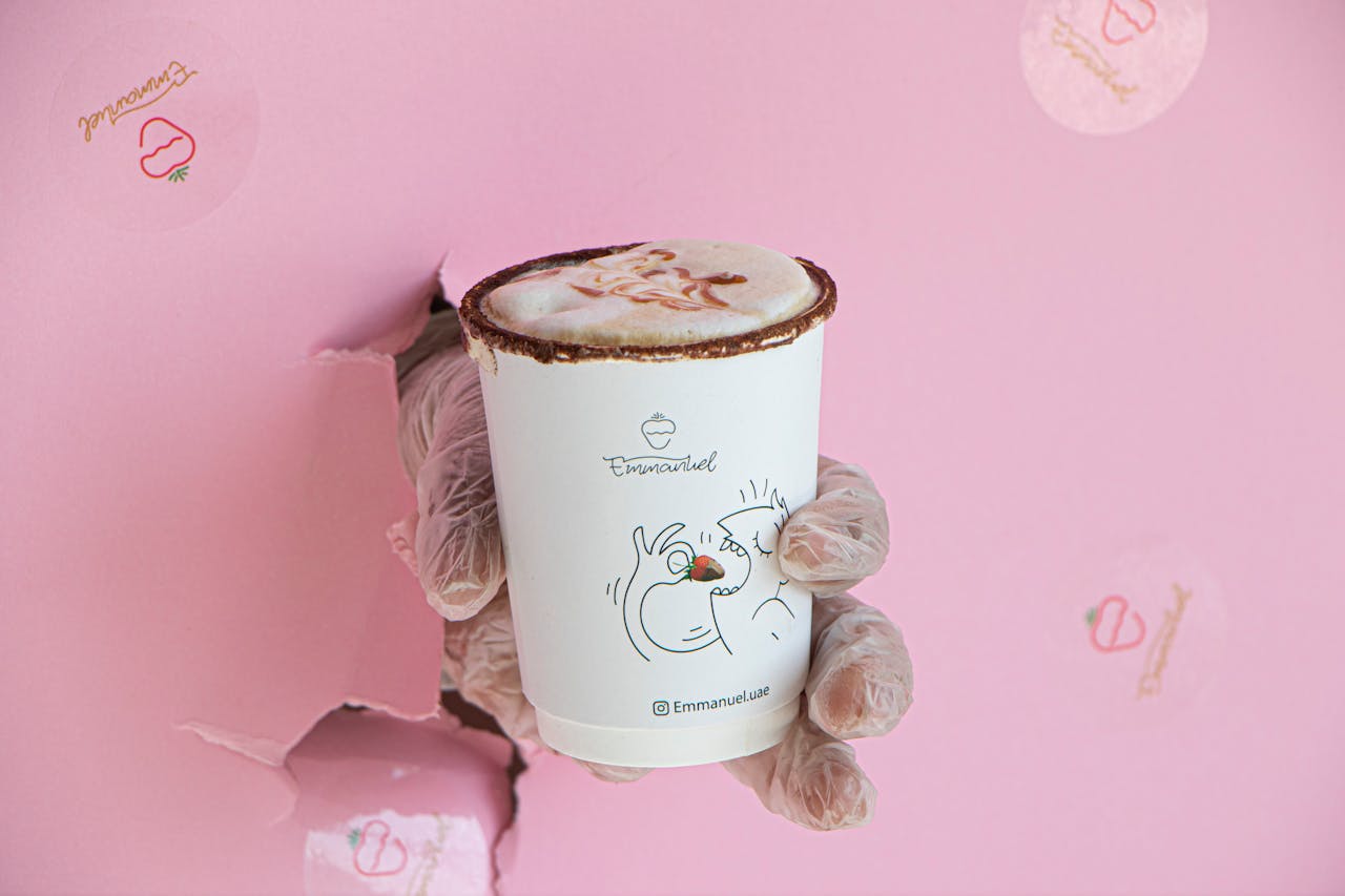 A hand in a glove holding a decorated coffee cup through a torn pink backdrop, creating a whimsical vibe.